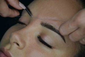 cara de mujer con ojos cerrados y extensiones de pestañas y manos de profesional haciendo una depilación de cejas a hilo
